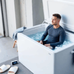 In an outdoor or garage setting, a large chest freezer is repurposed as a DIY ice bath, with its lid open to reveal clear water and floating ice. Nearby, a wooden step stool, a towel, a timer, and a pair of sliders create a practical and disciplined atmosphere, while a gender-neutral person sits calmly in the water, emphasizing the cold water immersion experience.